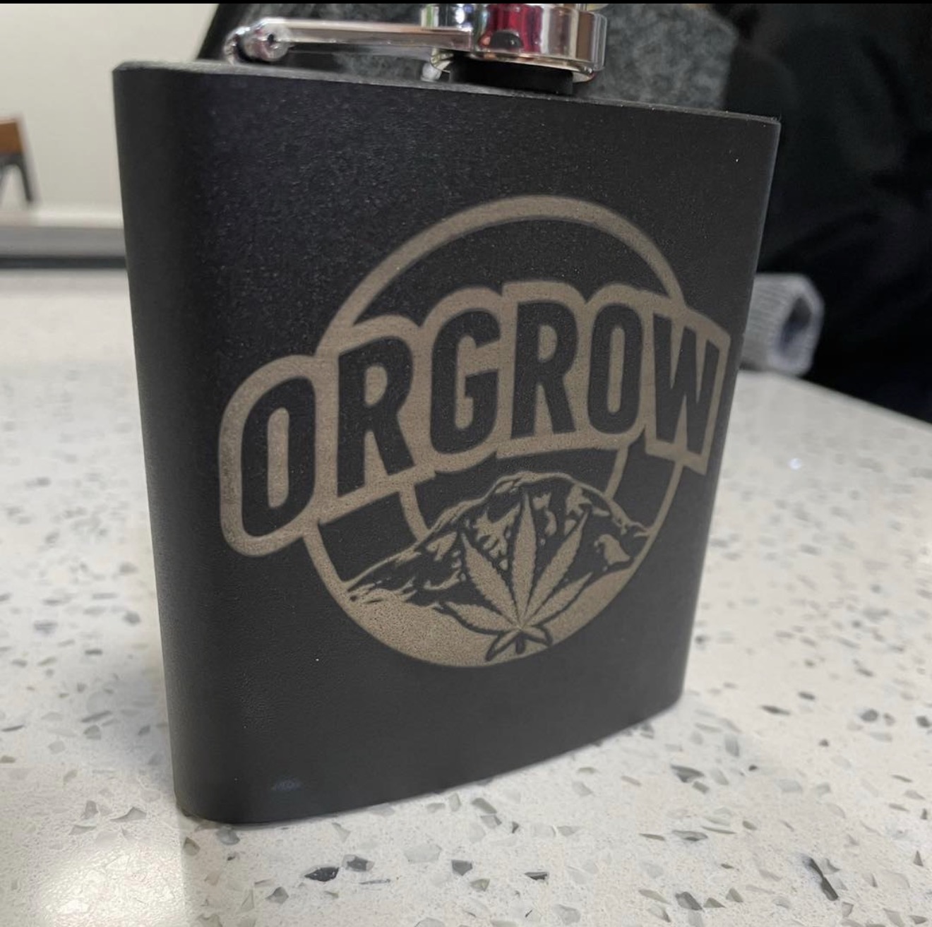 Black metal flask engraved with the OrGrow logo featuring a mountain and cannabis leaf.
